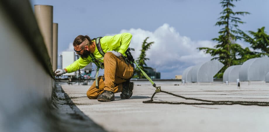 Bearded man repairs waterproofing on large commercial roof, kneeling on one knee attached to a safety rope
